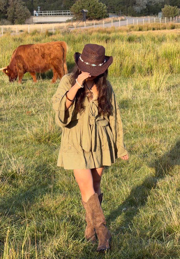 Una mujer con sombrero de vaquero, vestido corto y botas camina por un campo de hierba mientras una vaca marrón pasta detrás de ella.