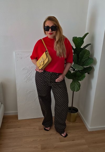 Mujer con cabello largo y rubio, llevando una blusa roja, pantalones negros con lunares, gafas de sol negras y sosteniendo un bolso amarillo en el interior cerca de una planta en maceta.
