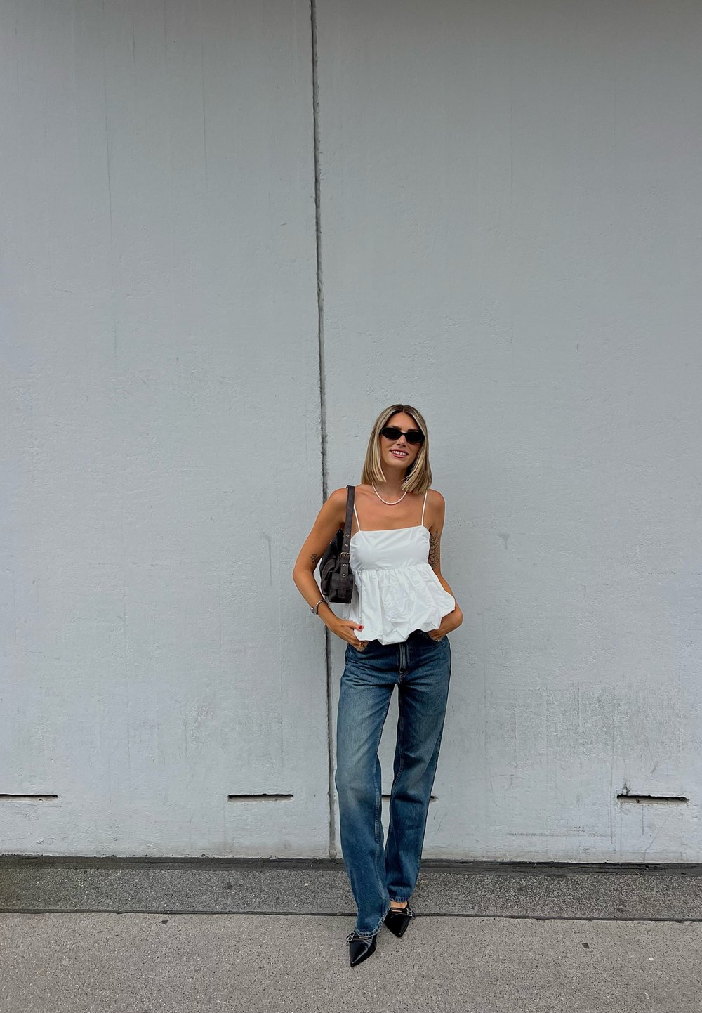 Top peplum branco com alças finas, combinado com jeans de ganga azuis. Acessórios incluem uma mala preta e sapatos de salto agudo pretos.