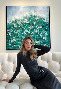 Γυναίκα με μακριά ξανθά μαλλιά και μαύρο φόρεμα καθισμένη σε λευκό καναπέ με καπιτονέ, με το χέρι πίσω από το κεφάλι, αφηρημένος πράσινος και λευκός πίνακας στον τοίχο πίσω της.