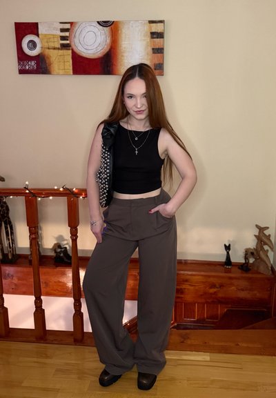 Top corto negro sin mangas y pantalones marrones de pierna ancha. Los accesorios incluyen collares en capas y un chaleco negro adornado con un lazo.