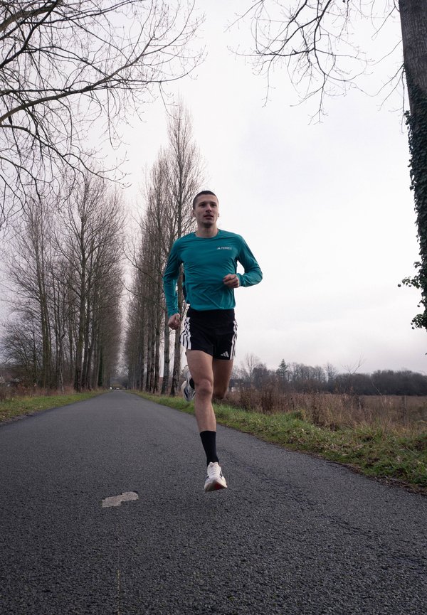 Homme courant sur une route bordée d'arbres, portant un haut à manches longues turquoise, un short noir, des chaussettes noires et des chaussures de course blanches par une journée nuageuse.