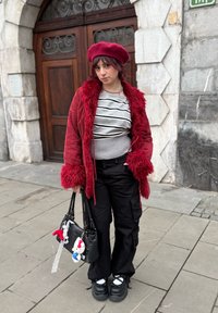 Veste en velours côtelé rouge avec garniture en fausse fourrure, pull rayé gris, pantalon cargo noir, chaussures plates-formes noires épaisses, et sac à main décoré.