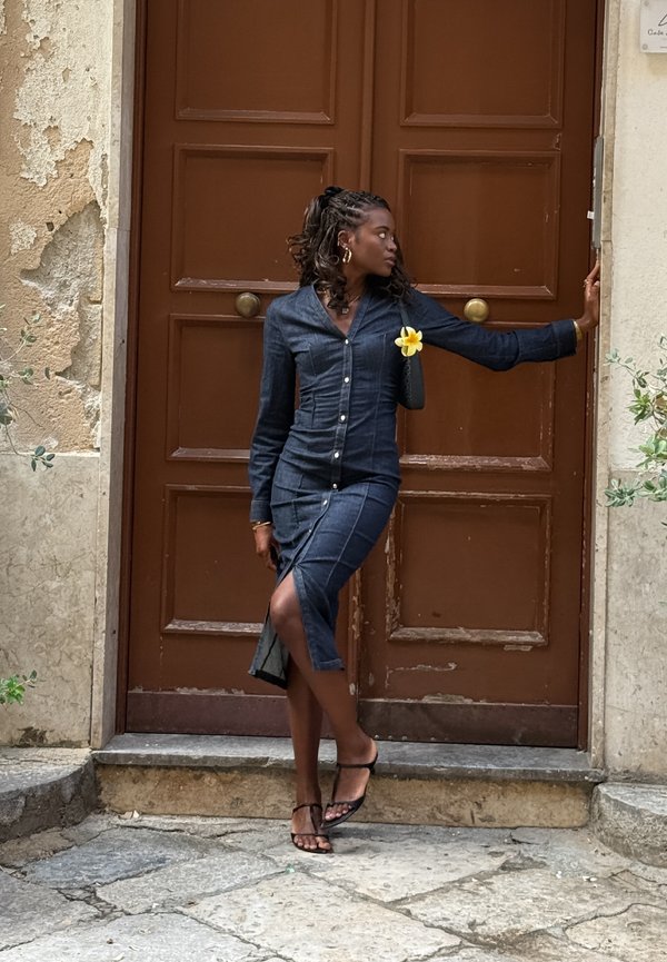Denim overhemddress in donkerblauw met een getailleerd silhouet, knoopdetails en een zijscheur. Geaccentueerd met een gele bloem en gestyled met sandalen.