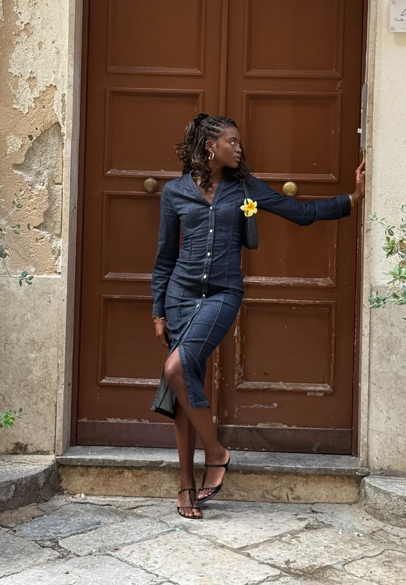 Jeansskjortklänning i mörkblått med figursydd siluett, knappdetaljer och sidospalt. Accenterad med en gul blomma och stylad med sandaler.