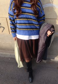 Pull en maille rayé bleu et jaune, chemise blanche en dessous, jupe marron avec ourlet en dentelle, bottes noires montant jusqu'aux genoux, tenant un sac de couleur crème.