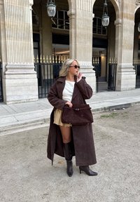 Lang brun semsket frakk med store slag, lagvis over en hvit topp og kremfarget skjørt. Brun h�åndveske og knelang svarte støvler fullfører antrekket.