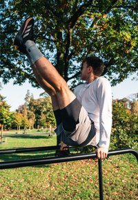 Juodi sportiniai batai su raudonais akcentais, pilki šortai ir balta ilgomis rankovėmis palaidinė. Asmuo atlieka kojų pakėlimą ant juodų paralelinių barų žaliame parke.