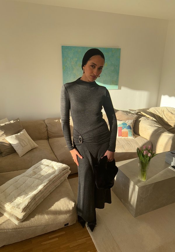 Žena v tmavých priliehavých šatách a šatke na hlave stojí v slnečnej obývačke s béžovou pohovkou, kvetmi vo váze a za ňou je abstraktný modrý obraz.