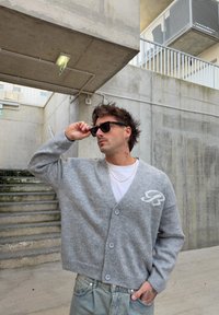 Cardigan in lana grigia con scollo a V e chiusura con bottoni, con dettaglio logo bianco sul petto. Indossato sopra una t-shirt bianca.