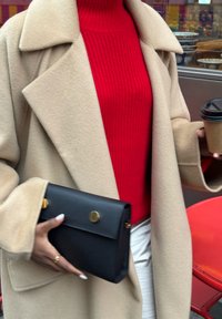 Beżowy płaszcz z szerokimi klapami na czerwonym prążkowanym swetrze, trzymający czarną kopertówkę z złotym zapięciem oraz kubek na wynos z kawą.