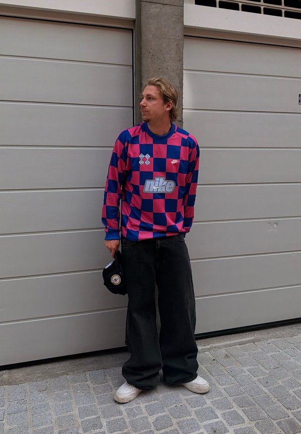 Mand i blå og pink ternet Nike sweatshirt og brede sorte bukser står og ser mod venstre, mens han holder en sort kasket foran en lukket garagedør.