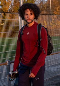 Mand med krøllet hår iført en rød langærmet skjorte og rygsæk, der holder et elektrisk skateboard nær en indhegnet sportsplads ved solnedgang.