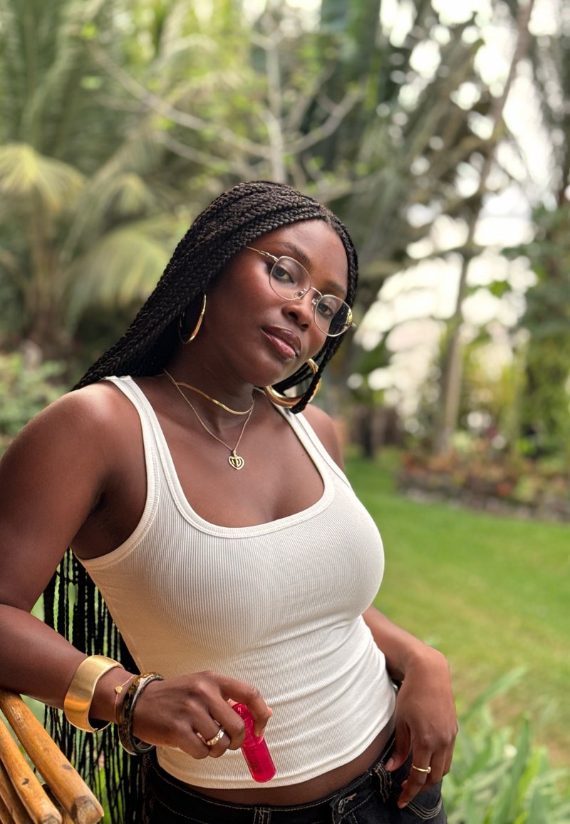 Femme aux longues nattes, lunettes et bijoux dorés, appuyée sur une rambarde en bois à l'extérieur, tenant une petite bouteille rouge sur un fond de jardin vert.