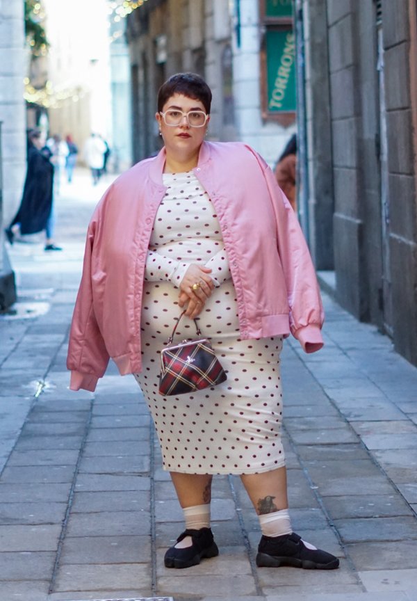 Rövid hajú személy fehér pöttyös ruhában és rózsaszín dzsekiben, kockás kézitáskát tartva, egy kőkőutcán áll.