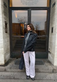 Veste en cuir noire, col roulé rayé gris et blanc, pantalon large rose pâle, sac noir, bottines à talons carrés, debout près de la porte.