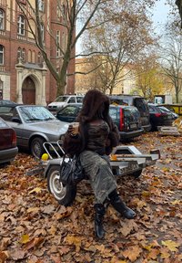 Personne en pantalon camouflage et veste à capuche en fourrure tenant une boisson, assise sur une petite remorque au milieu des feuilles d'automne tombées dans un parking urbain.
