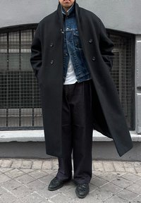 Άνδρας που φοράει μαύρο μακρύ παλτό σε στρώσεις πάνω από μπλε τζιν μπουφάν, στέκεται σε πεζοδρόμιο με κυβόλιθους μπροστά από παράθυρο με μεταλλικά κάγκελα.
