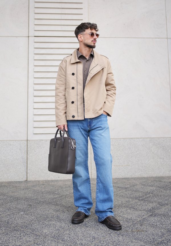 Homme aux cheveux courts et foncés avec une barbe, debout portant des lunettes de soleil, une veste beige, une chemise marron, un jean bleu, des chaussures noires, tenant une mallette en cuir noire.
