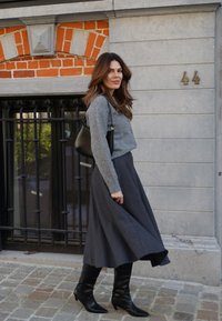 Grauer Pullover, langer dunkler Rock und schwarze kniehohe Stiefel. Mit strukturierter Stoffqualität und fließendem Design. Schwarze Handtasche über der Schulter. Ziegelsteinmauer im Hintergrund.