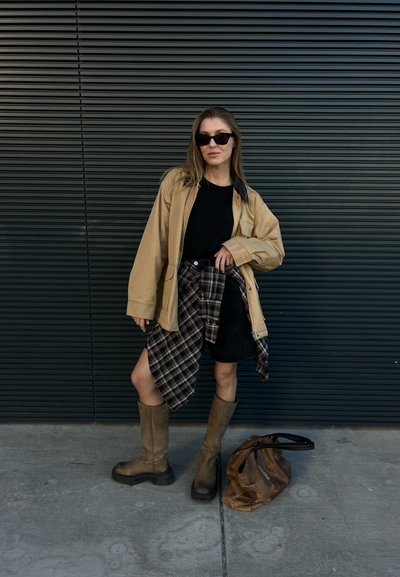 Chaqueta oversize marrón, camiseta negra, una camisa de franela a cuadros atada a la cintura, falda negra, botas marrones hasta la rodilla y un bolso marrón en el suelo.