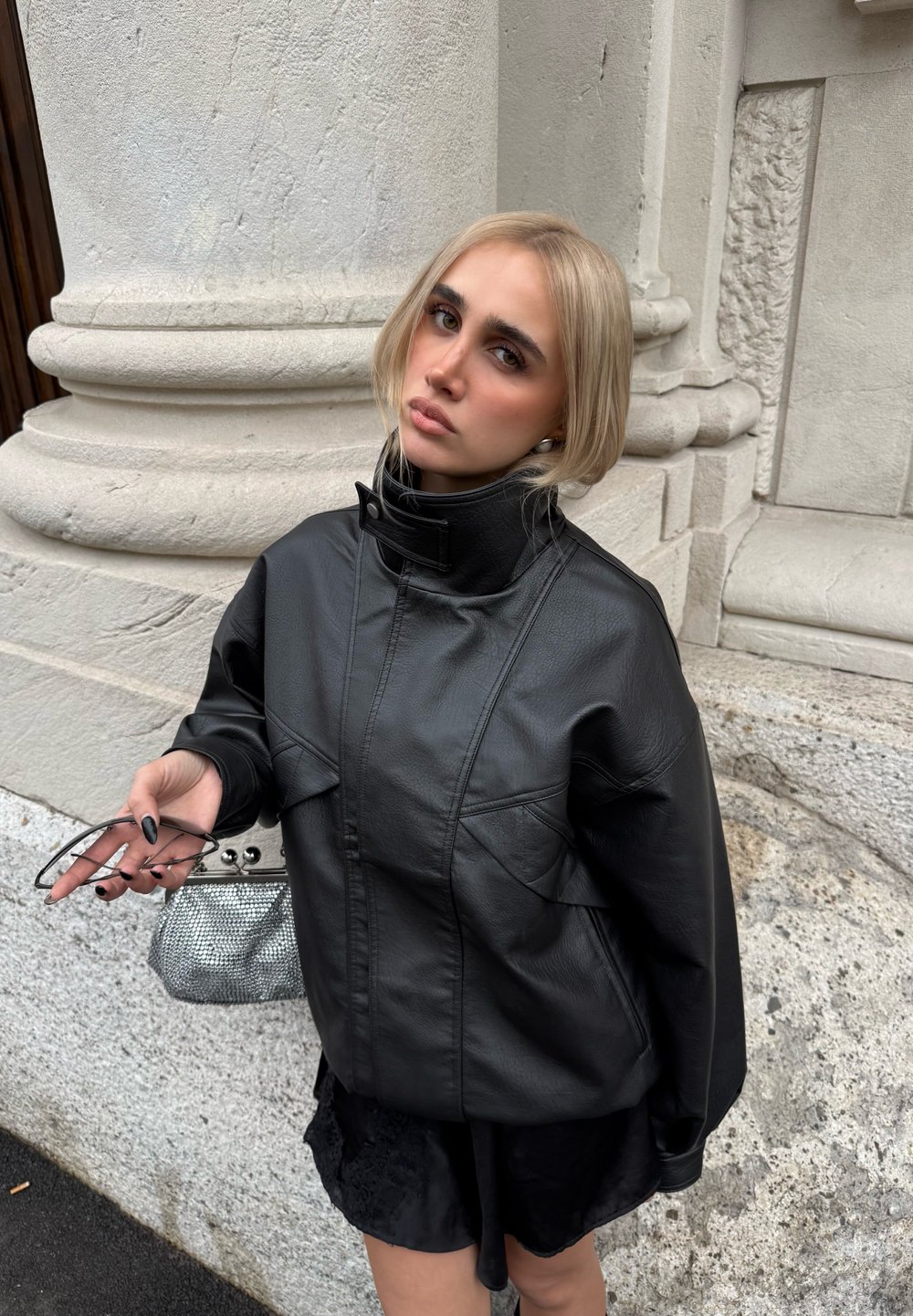 Schwarze, übergroße Lederjacke mit hohem Kragen, minimalen Nähten und Taschen. In der Hand gehalten: ein Netzbeutel und Sonnenbrillen mit Metallakzenten.