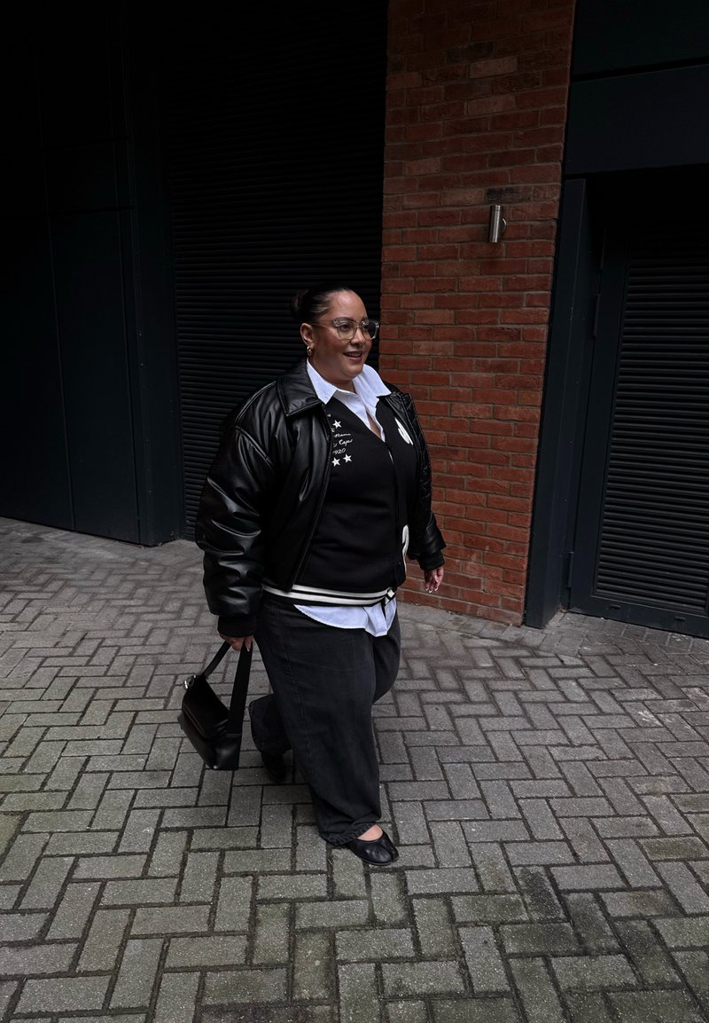 Femme portant des lunettes, veste en cuir noire, pull noir, chemise blanche, pantalon noir et chaussures plates, marchant sur un sol pavé près d'un mur en briques, portant un sac noir.