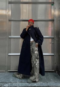 Pitkä laivastonsininen trenssitakki, kuvioidut maastokuvioiset fingustinhousut, vihreät paksupohjaiset kengät ja punainen neulottu päähuivi. Kirkkaat silmälasit ja näyttävät korut.