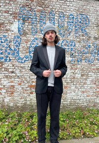 Černý blazer se saténovými klopami přes šedé tričko, v kombinaci s černými kalhotami. Postava stojí před cihlovou zdí s modře natřeným textem.