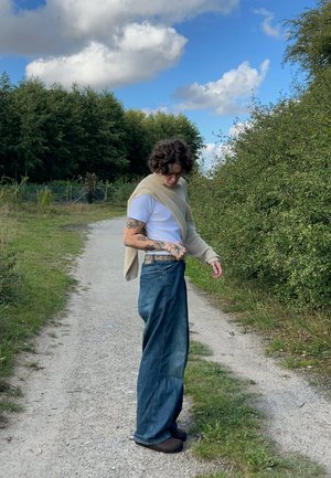 Jeans de mezclilla azul de pierna ancha con cintura alta y dobladillos deshilachados, combinados con una camiseta blanca y un suéter de punto beige claro sobre los hombros.