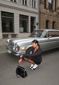 Černá kabelka s bílými akcenty, sedící postava v černém pruhovaném tričku a sukni, stříbrný vintage automobil s chromovými detaily v městském prostředí.