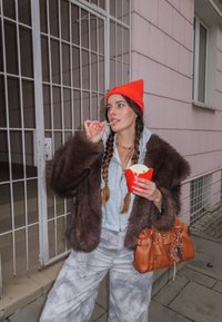 Brązowy płaszcz z futerkiem syntetycznym, szara bluza z kapturem, wzorzyste spodnie dresowe, pomarańczowa czapka, trzyma czerwoną paczkę frytek, brązowa torebka, plener z kratkowanym tłem.