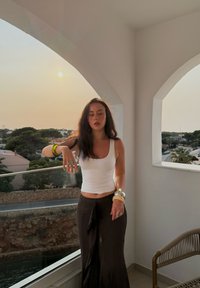 Weißes Tanktop und brauter, fließender Rock. Mehrere Armbänder an einem Handgelenk. Der Hintergrund zeigt einen Balkon mit einem Bogen und Sonnenuntergang.