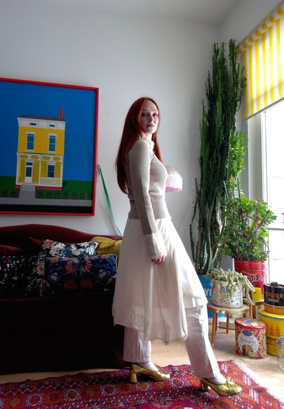 Joven con pelo largo y rojo que lleva un suéter beige, falda blanca sobre pantalones y tacones dorados, de pie en una sala de estar colorida con plantas y obras de arte.