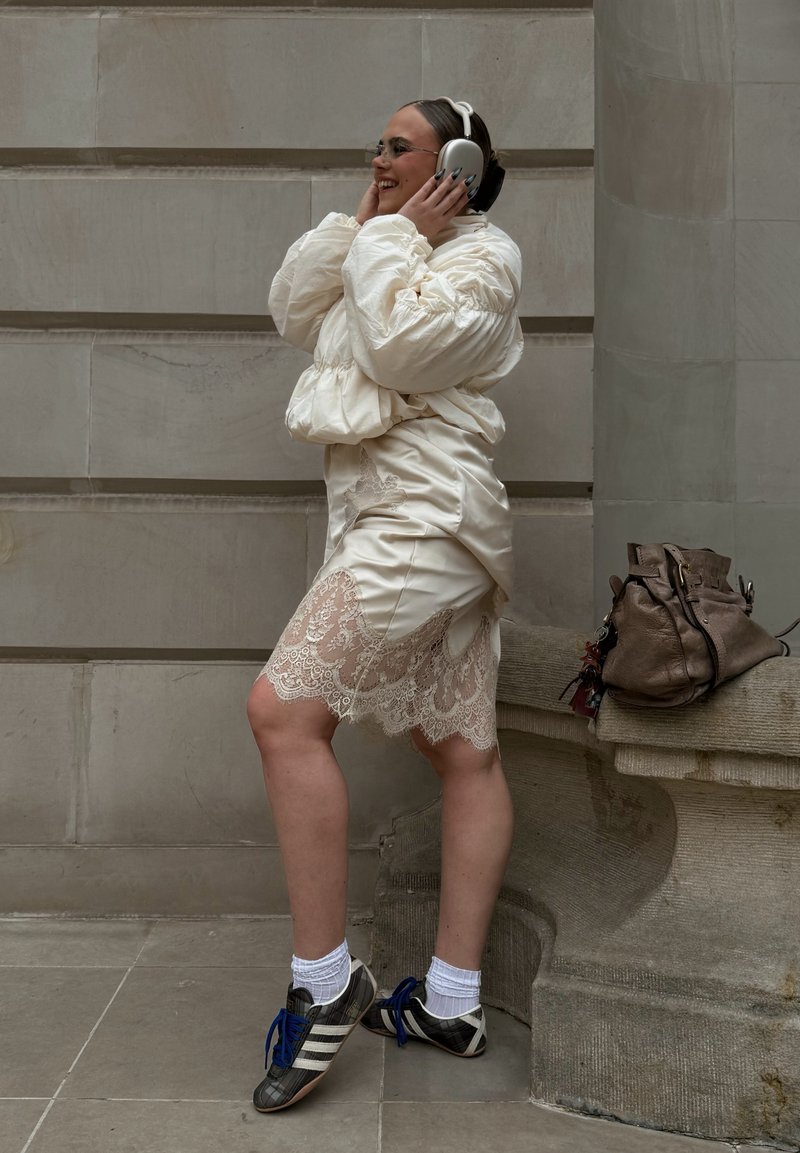 Femme en robe blanche en dentelle et veste gonflée, portant un casque, debout sur une jambe près d'un banc en pierre avec un sac marron posé dessus.