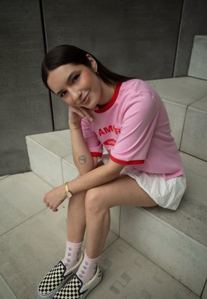 Camiseta rosa con acentos rojos y texto "SOY", falda blanca, zapatos de lona a rayas en blanco y negro, y calcetas con patrón floral. Sentada en unos escalones.