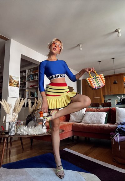 Camiseta de manga larga azul corta con el logo "PANTSCOU", falda plisada de rayas amarillas, bolso de mano tejido colorido y zapatos con estampado. Ambiente interior.