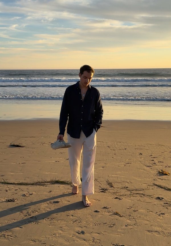 Junger Mann, der barfuß bei Sonnenuntergang am sandigen Strand entlanggeht, hält Schuhe in einer Hand, im Hintergrund Ozeanwellen und Himmel.