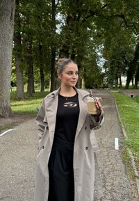 Ung kvinde i beige trenchcoat og sort outfit, der holder en iset kaffe og solbriller, mens hun står på en træbelagt parkvej.