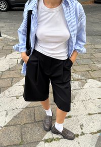 Camisa de botão às riscas azuis e brancas, top branco canelado, calções pretos plissados, meias brancas e mocassins castanhos numa rua de paralelepípedos.