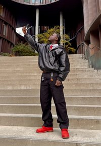 Czarna skórzana kurtka z kapturem, biała koszula z kołnierzykiem, czerwony krawat, czarne spodnie i czerwone buty. Trzyma smartfon, stoi na betonowych schodach.