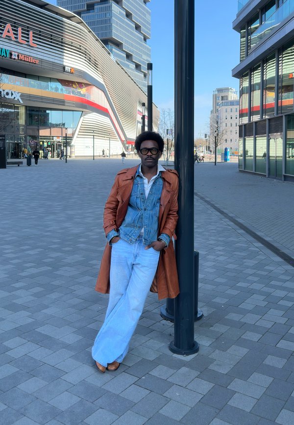 Mand iført brun læderjakke og denimjakke læner sig op ad en gadelygte på en bred urban fodgængerplads nær moderne bygninger.