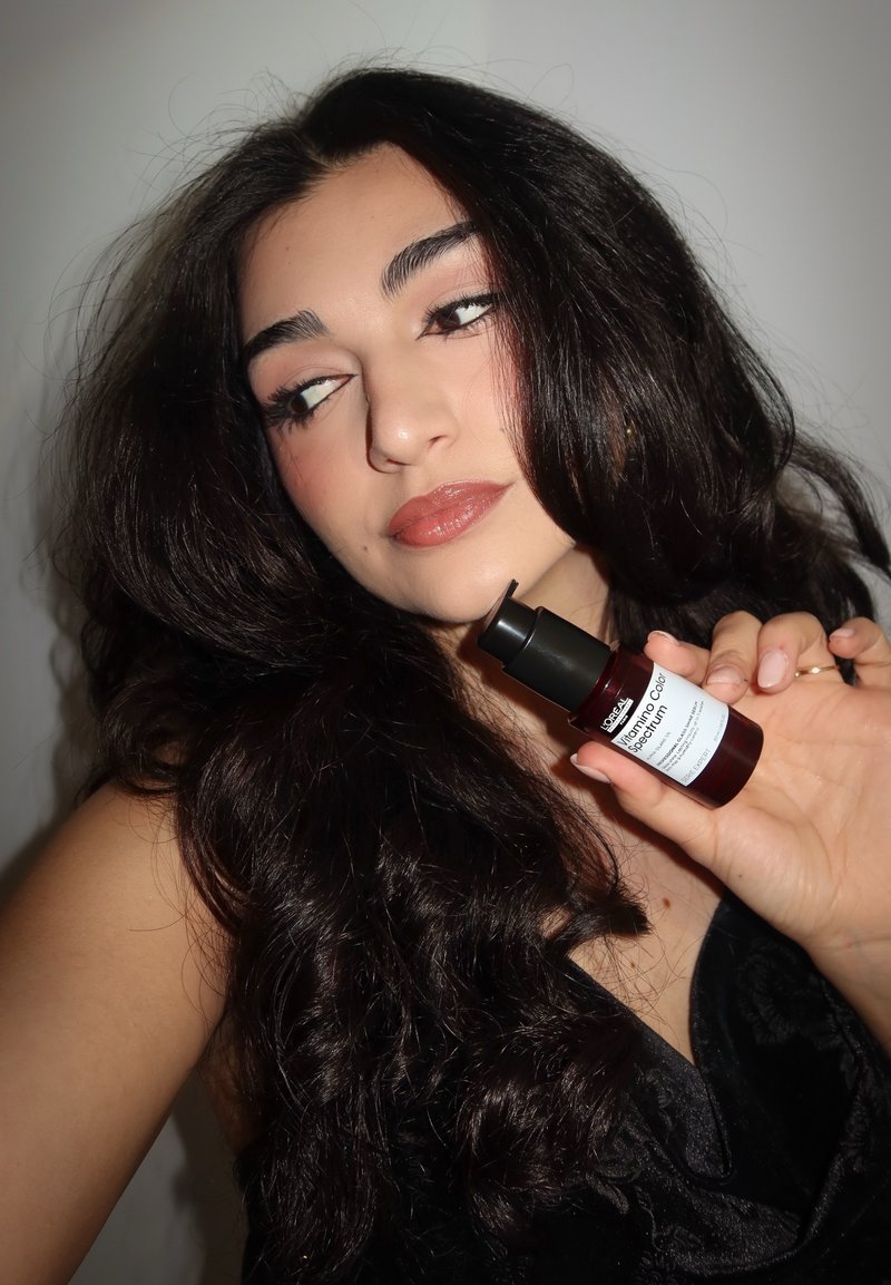 Femme aux longs cheveux bruns tenant une bouteille rouge foncé de sérum L'Oréal Vitamin Color Spectrum près de son visage, regardant sur le côté.