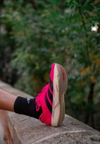Perna estendida sobre um muro de pedra, calçando um ténis de corrida rosa vivo e preto com uma meia atlética preta de meio cano, contra um fundo desfocado de folhagem verde.