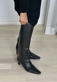 Femme portant des bottes en cuir noires à bout pointu jusqu'aux genoux et une jupe côtelée noire, debout sur un sol en bois clair.