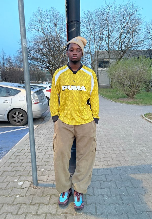 Camisola amarela de manga longa da Puma com um padrão texturizado, detalhes pretos, calças cargo bege e sapatilhas multicoloridas. Cenário urbano com árvores.