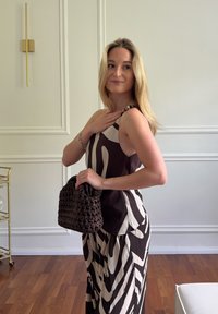 Mujer con cabello largo y rubio, que lleva un vestido con estampado en blanco y negro y sostiene un bolso tejido oscuro en una habitación luminosa con suelo de madera.