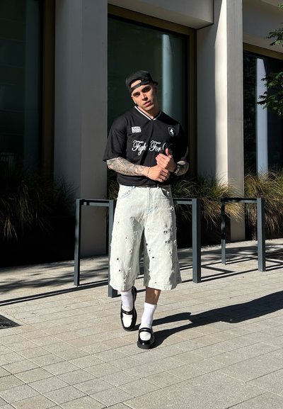 Camiseta negra con detalles blancos, shorts de mezclilla claros y desgastados, calcetines blancos, zapatos negros; la persona está de pie frente a un edificio moderno.