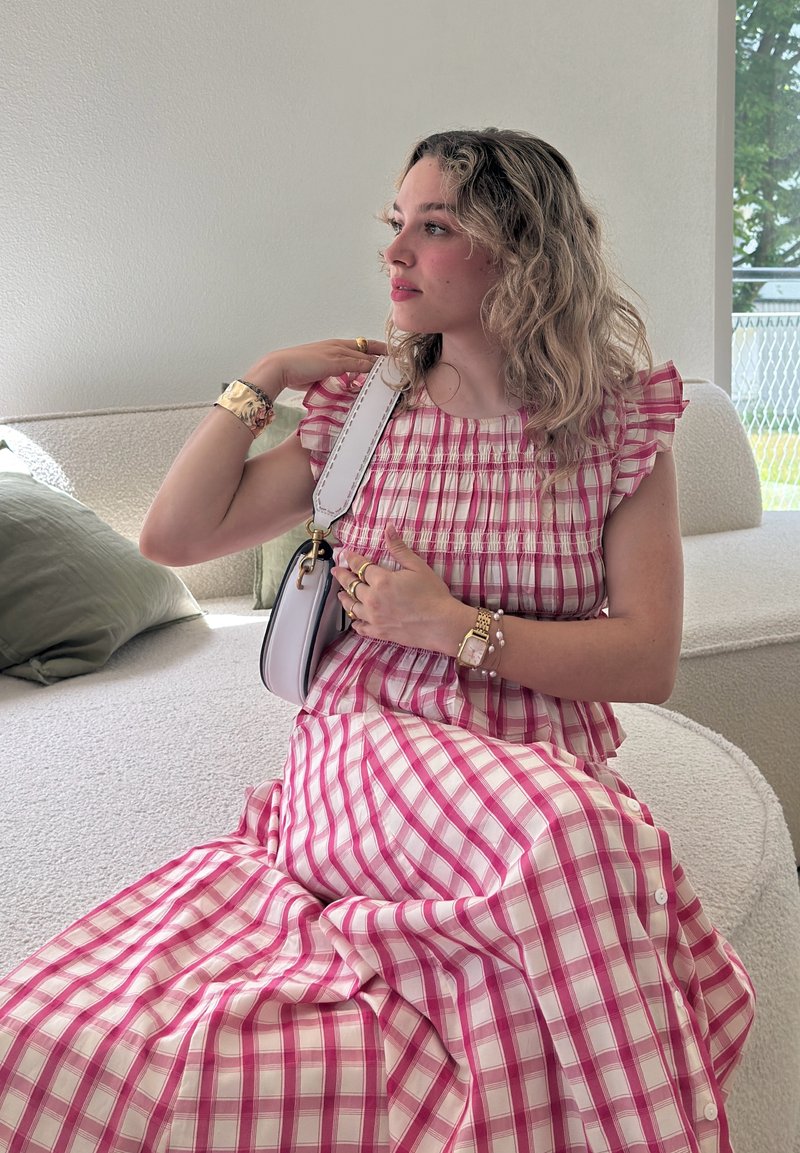 Jeune femme aux cheveux ondulés portant une robe rose à carreaux, assise sur un canapé clair, tenant une bandoulière de sac blanc sur son épaule.