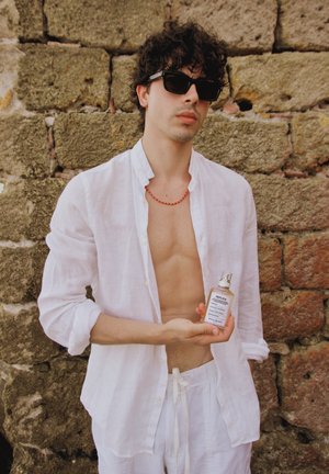 Camisa y pantalones de lino blancos, sosteniendo un frasco de perfume con tapa plateada. Pared de piedra texturizada de fondo. Gafas de sol y collar rojo.